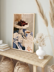 Watercolour still life print of cherries on a cake stand and a teacup on a blue tablecloth, in an alternate framed mockup
