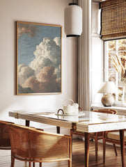 Detailed painting of voluminous white and reddish-brown cumulus clouds against a pale blue sky, in a styled room mockup