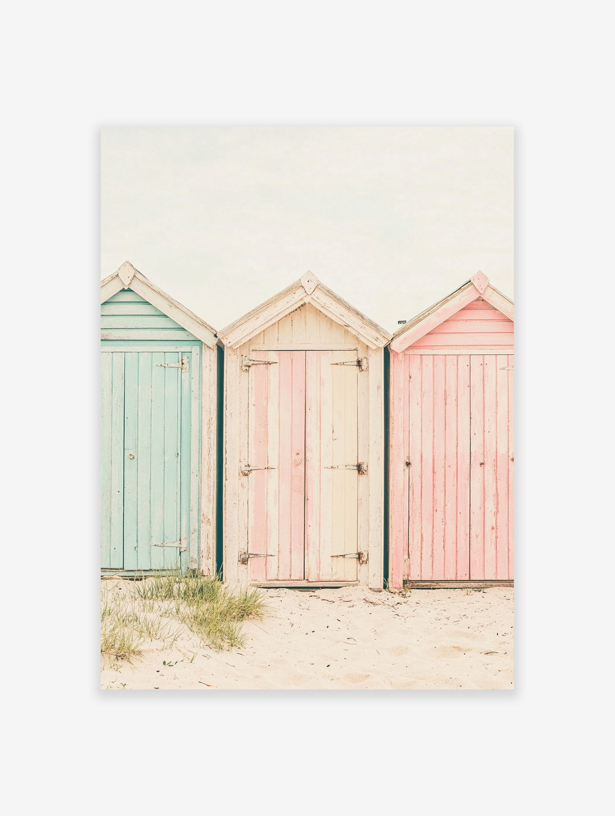 Photography poster of three weathered beach huts in pastel blue, pink, and striped cream on a sandy beach.