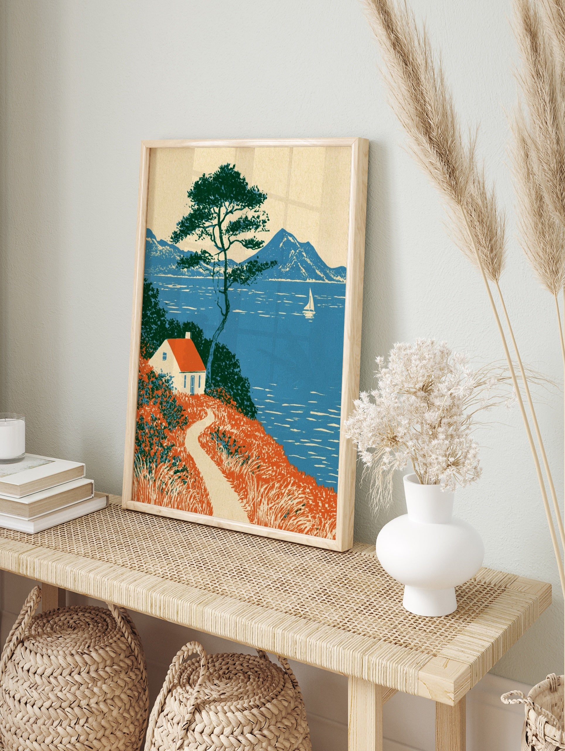Coastal cottage wall art with red roof house, pine tree, and sailboat on blue sea landscape, in an alternate framed mockup
