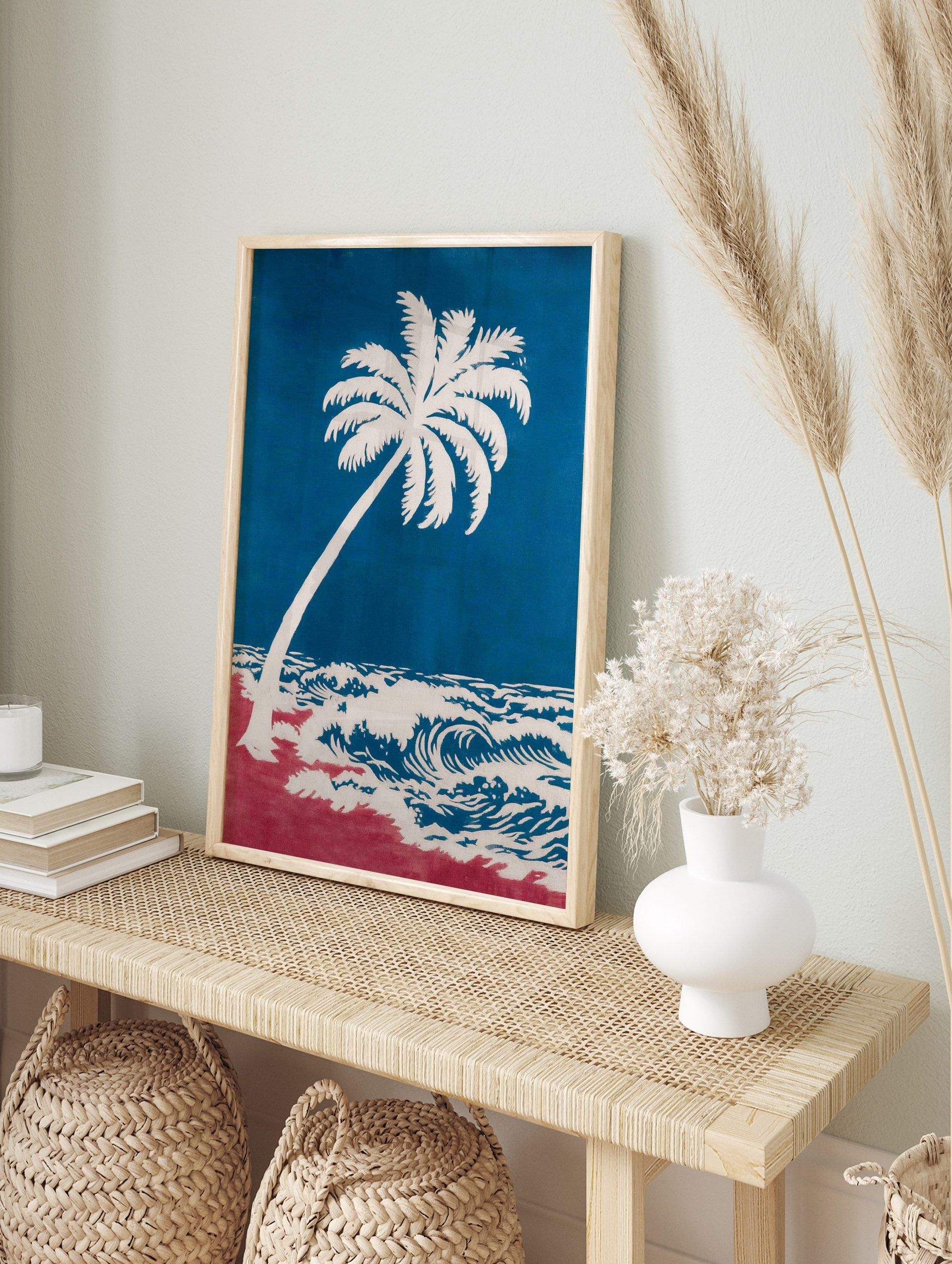 Coastal art print of a white palm tree leaning over blue ocean waves and a red beach, in an alternate framed mockup