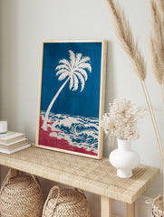 Coastal art print of a white palm tree leaning over blue ocean waves and a red beach, in an alternate framed mockup
