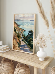 Watercolor-style print of a winding road descending through grassy hills towards a calm, in an alternate framed mockup