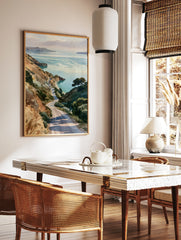 Watercolor-style print of a winding road descending through grassy hills towards a calm coastal bay, in a styled room mockup