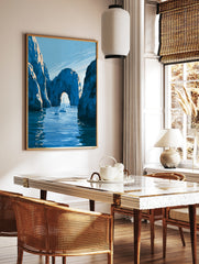 Illustrative coastal wall art print of a natural stone archway over blue water with sailboats, in a styled room mockup