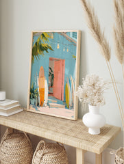 Painterly art print of a light blue building, pink door, and orange, white, yellow surfboards, in an alternate framed mockup