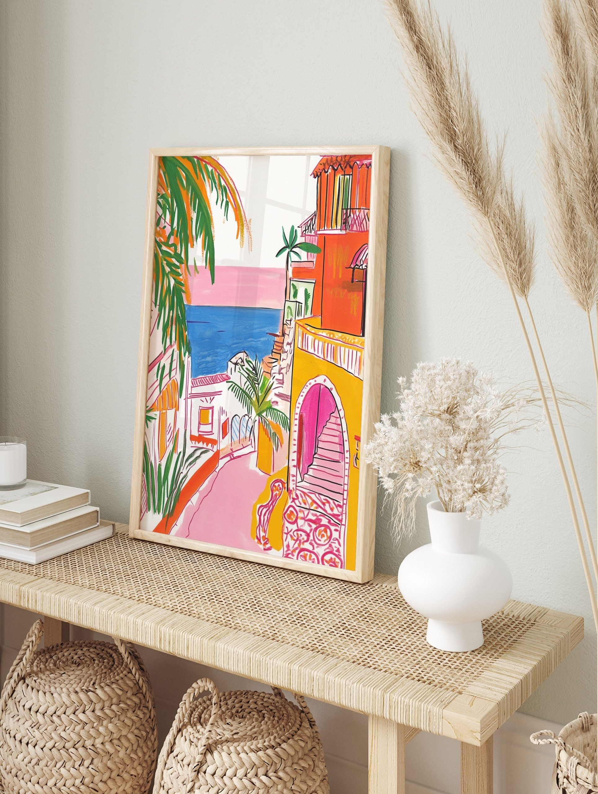 Illustrative coastal town scene with orange buildings, pink street, blue sea, and green palm, in an alternate framed mockup