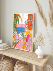 Illustrative coastal town scene with orange buildings, pink street, blue sea, and green palm, in an alternate framed mockup