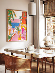 Illustrative coastal town scene with orange buildings, pink street, blue sea, and green palm trees, in a styled room mockup