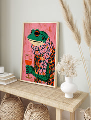 Illustrative colourful frog in floral shirt and scarf holding a wine glass on pink background, in an alternate framed mockup