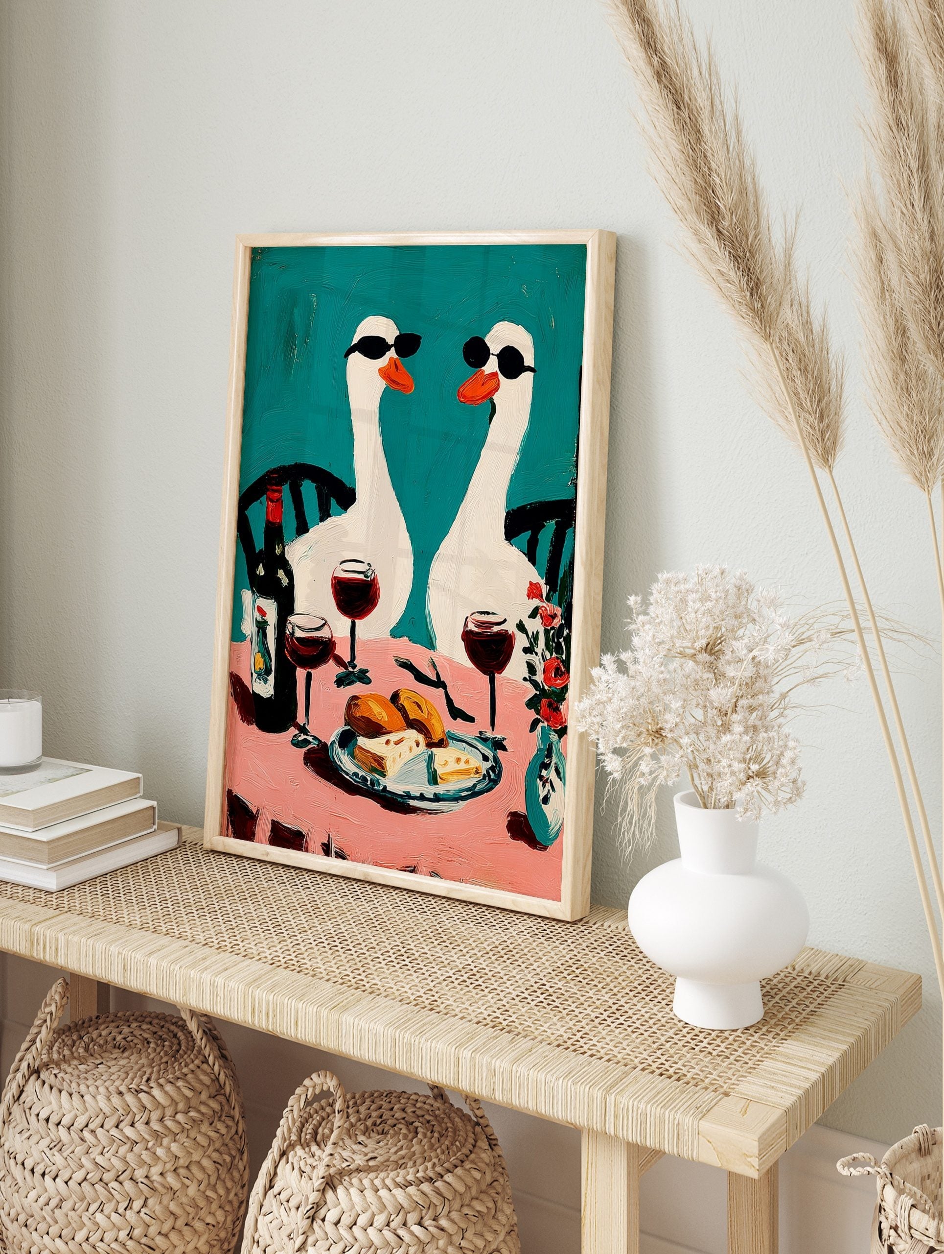 Two white geese in sunglasses at a pink table with wine and food, against a teal background, in an alternate framed mockup
