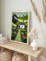 Linocut print of a cottage garden with a white building, cobbled path, raised beds, laundry, in an alternate framed mockup