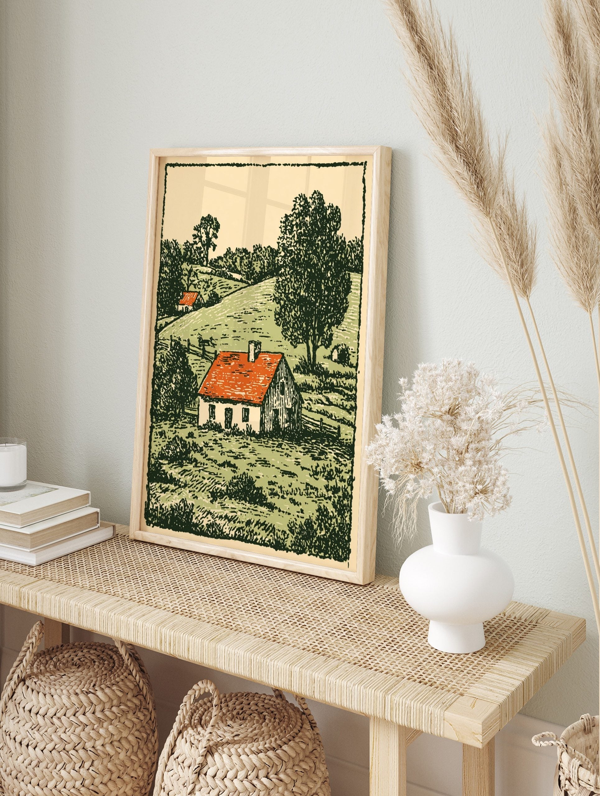 Woodcut style print of a white farmhouse with a red roof in a green, hilly countryside, in an alternate framed mockup