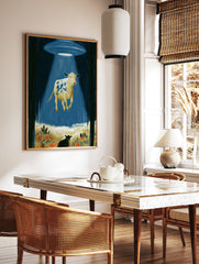 Surreal wall art print of a spotted cow levitating in a bright blue UFO tractor beam within a dark, in a styled room mockup
