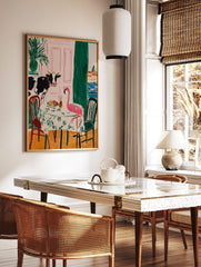 Illustrative wall art of a cow and a pink flamingo dining at a table, with a pink door and cityscape, in a styled room mockup