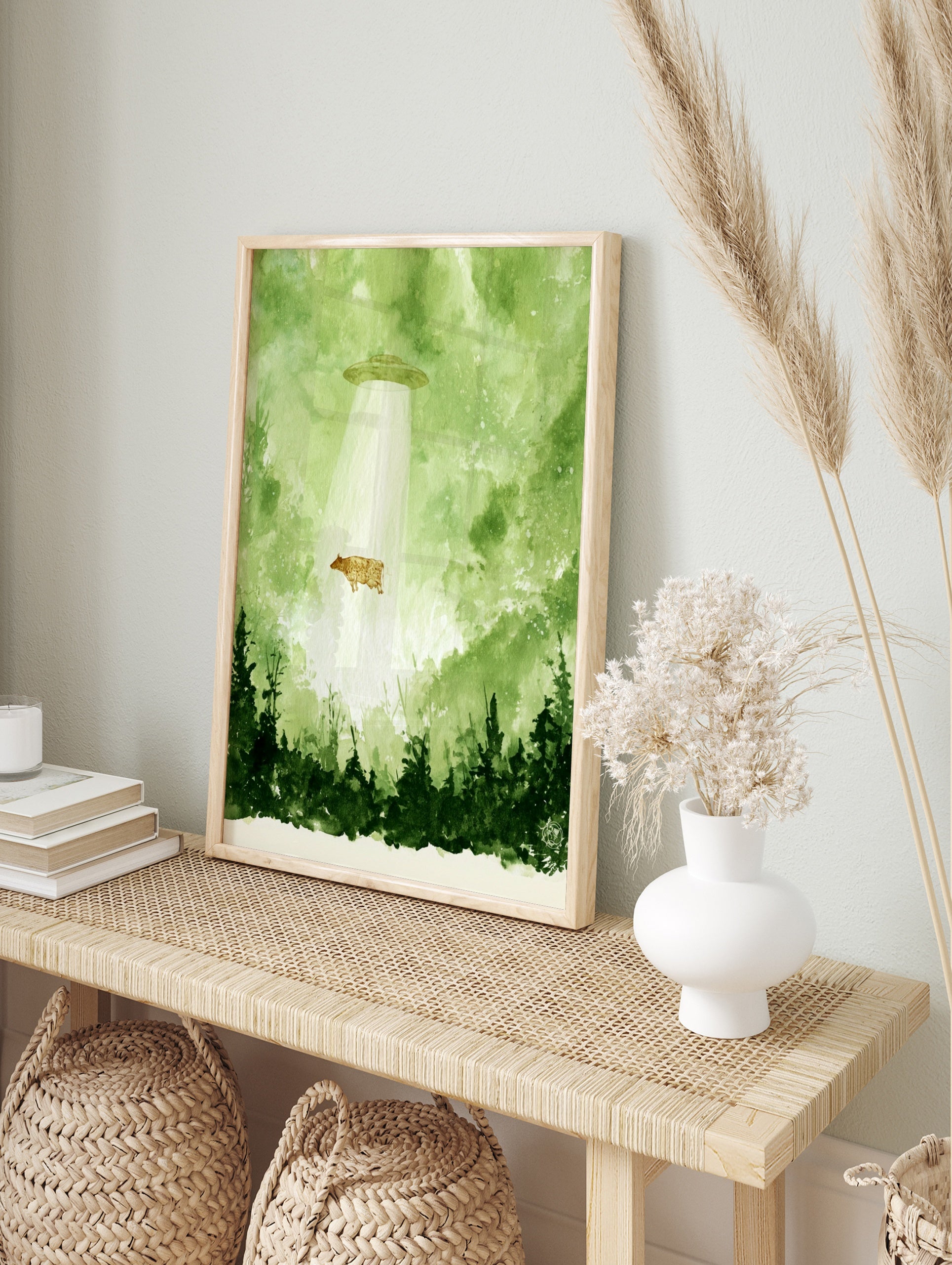 Watercolour illustration of a cow being abducted by a UFO in a green forest setting, in an alternate framed mockup