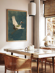 Illustrative art print of a white crane flying with a green frog on its back, over a deep blue, in a styled room mockup