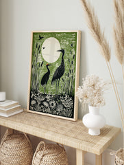 Green linocut print of two black cranes in a marsh with cattails and a large white moon in, in an alternate framed mockup