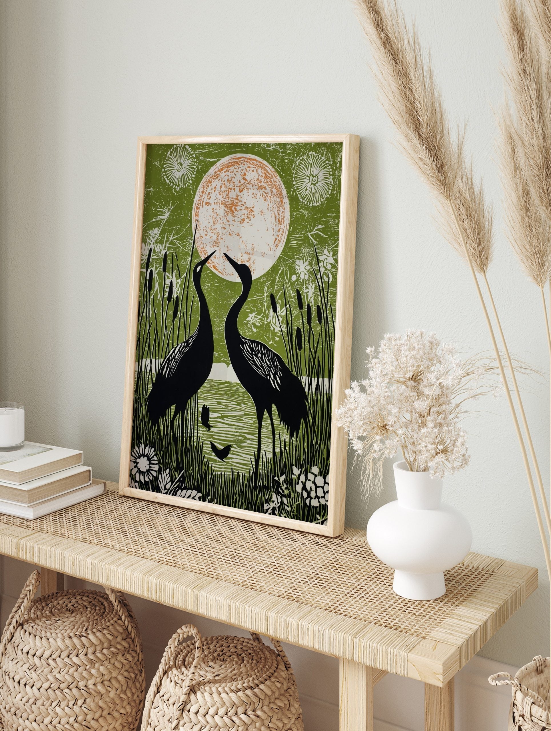Two black cranes facing a large orange and white moon, surrounded by green reeds and water in, in an alternate framed mockup