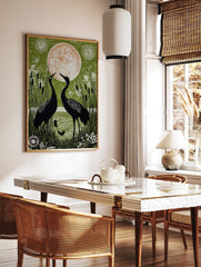 Two black cranes facing a large orange and white moon, surrounded by green reeds and water in an, in a styled room mockup