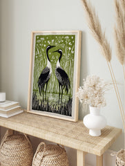 Linocut-style print of two black and white cranes in a green marsh with white hearts and reeds, in an alternate framed mockup