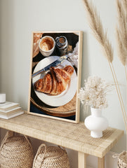 Watercolor painting of a croissant on a plate with coffee and a butter knife on a wooden, in an alternate framed mockup
