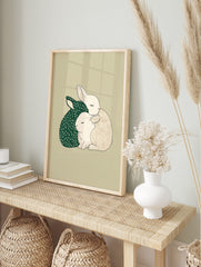 Illustrative print of two cuddling rabbits, one dark green with stars, one beige with a grid, in an alternate framed mockup