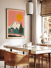 Cycling wall art illustration of a person riding a bicycle on a path with snowy mountains, a large orange sun, and a pine tree. - Image 2
