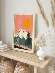 Cycling wall art illustration of a person riding a bicycle on a path with snowy mountains, a large orange sun, and a pine tree. - Image 3