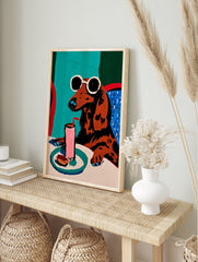 Illustration of a brown dachshund with sunglasses drinking a pink beverage at a table, in an alternate framed mockup