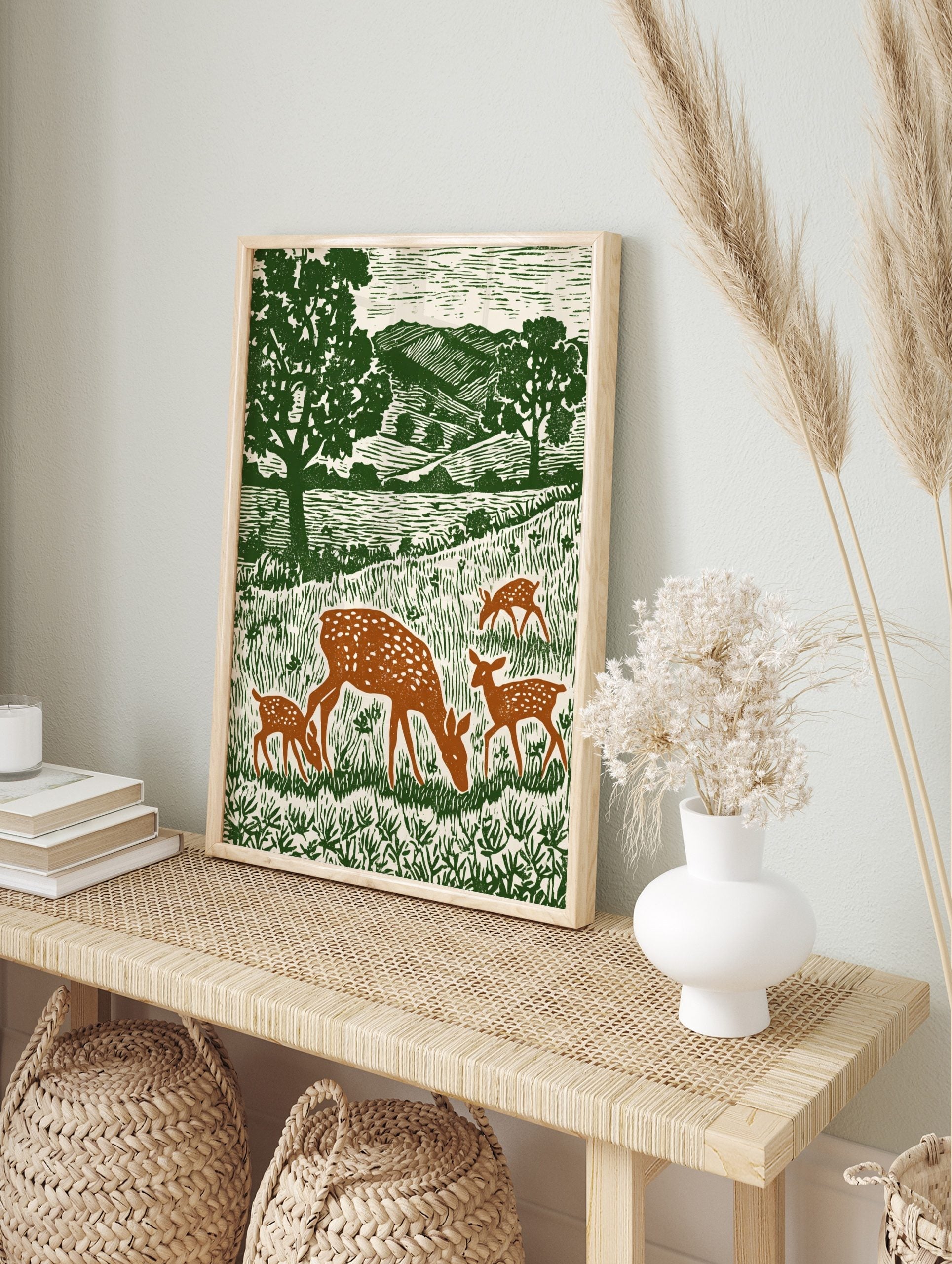 Woodcut style print of a spotted doe and three fawns grazing in a green meadow with trees and, in an alternate framed mockup