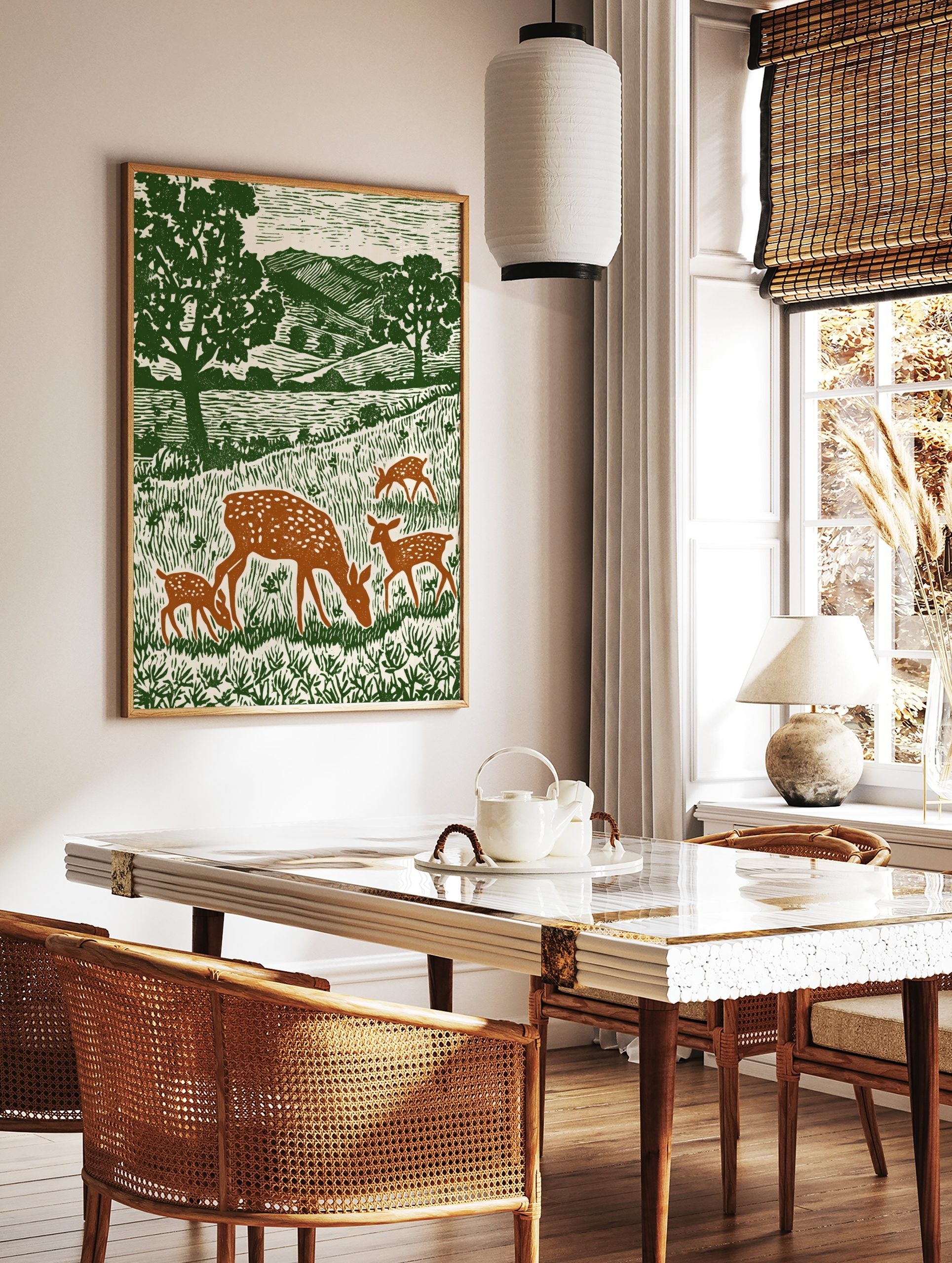 Woodcut style print of a spotted doe and three fawns grazing in a green meadow with trees and hills, in a styled room mockup