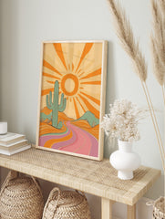Retro desert landscape art print with a large teal cactus, orange sun, and a winding pink and, in an alternate framed mockup