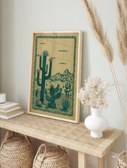 Illustrative desert landscape print with teal green saguaro and prickly pear cacti against a, in an alternate framed mockup