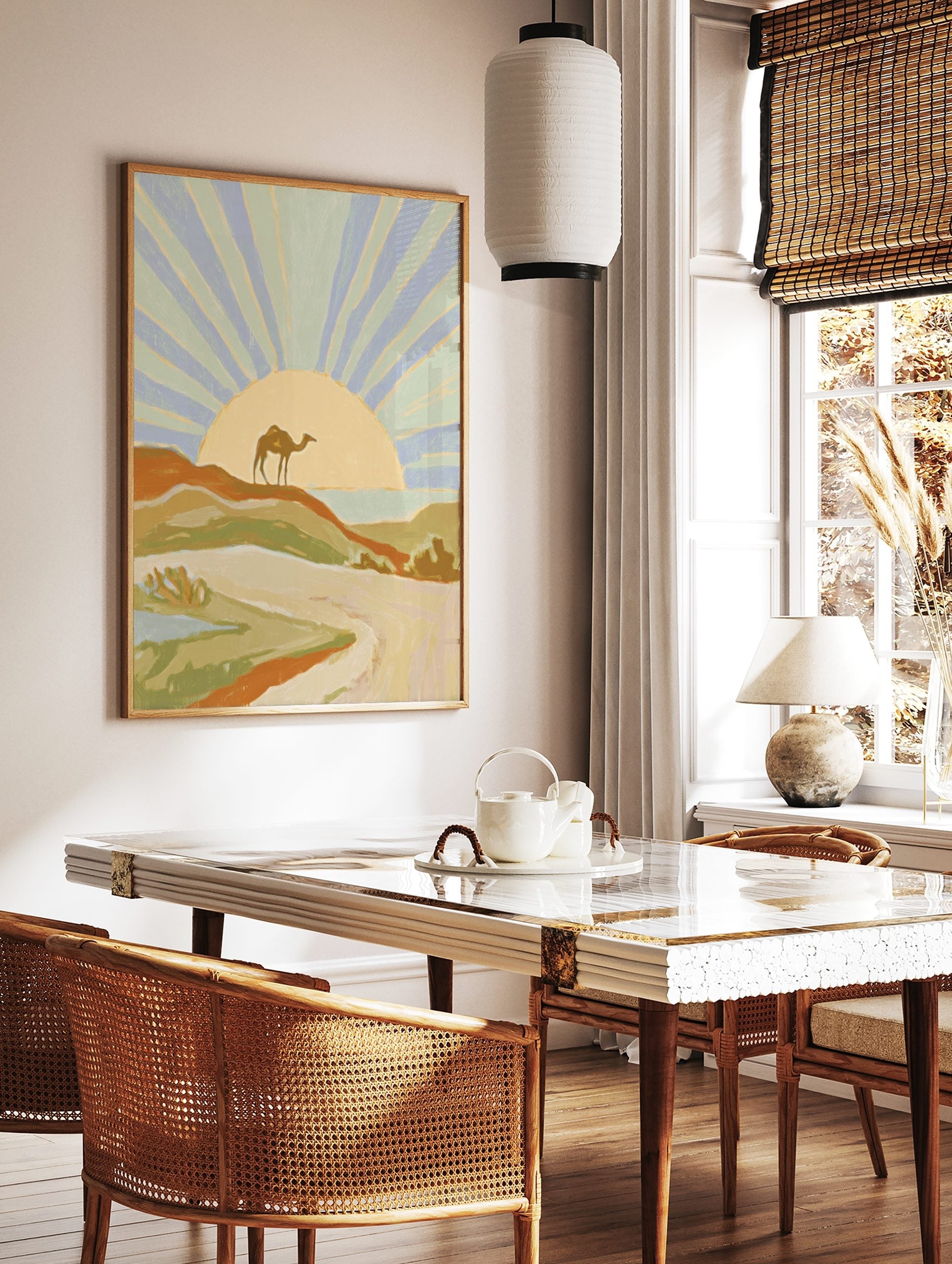 Illustrative desert landscape art print with a camel on a dune, radiating sunset, and distant sea, in a styled room mockup