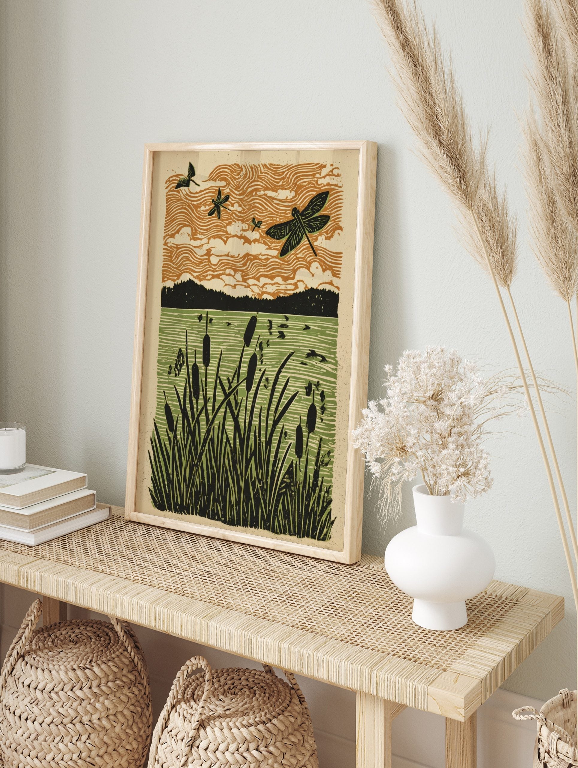 Linocut style art print of dragonflies flying over cattails in a marshland with an orange sky, in an alternate framed mockup