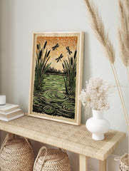 Illustrative woodcut style print of cattails, green water with ripples, and three dragonflies, in an alternate framed mockup
