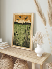 Linocut-style print of black dragonflies flying over a green pond with cattails and trees, in an alternate framed mockup