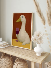 An illustrative art print of a white duck holding a glass of red wine against a warm red, in an alternate framed mockup