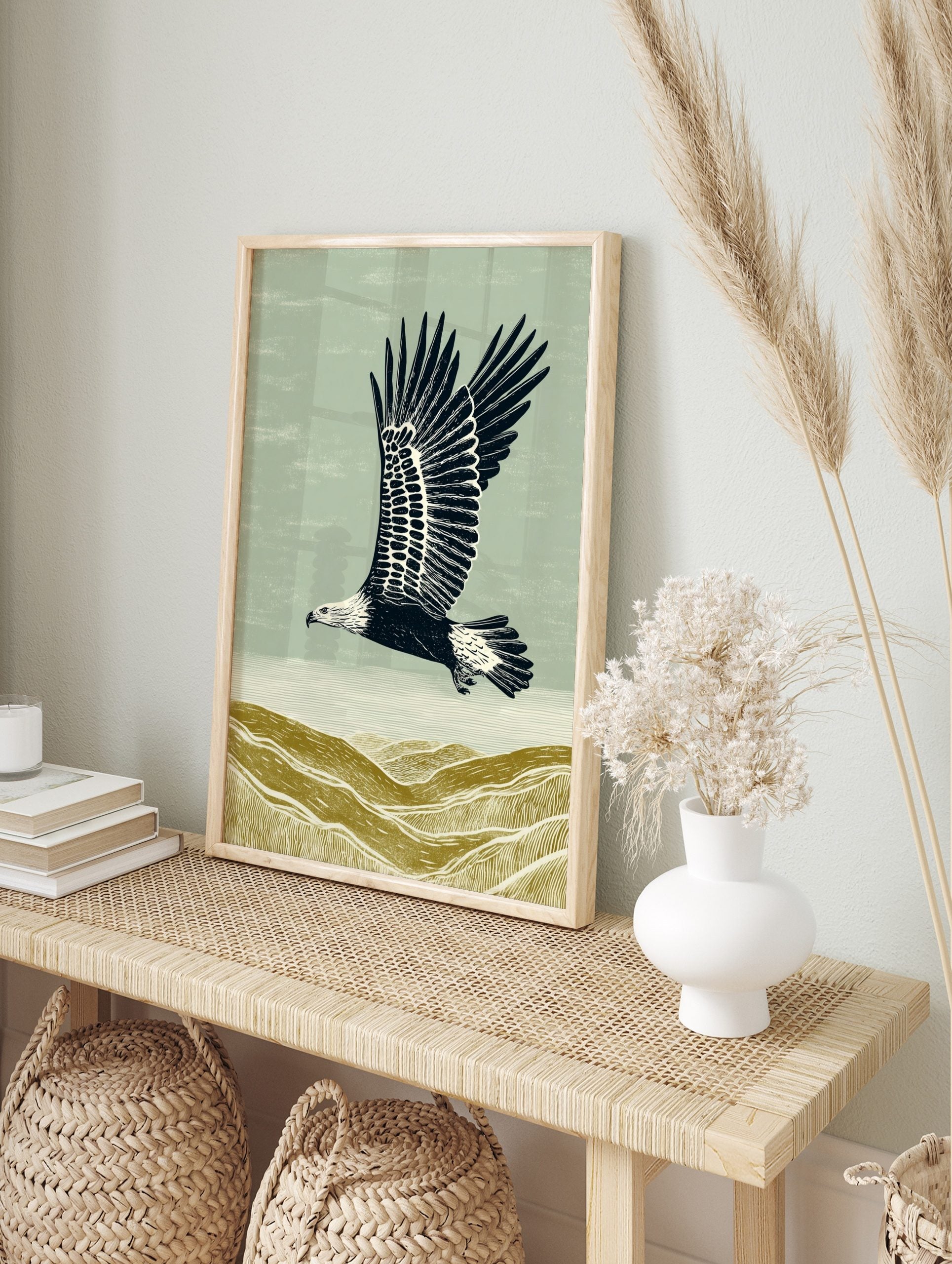 Illustrative linocut style print of an eagle flying over textured olive green hills under a, in an alternate framed mockup