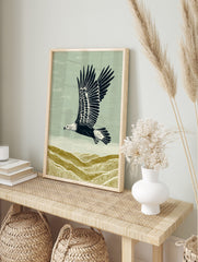 Illustrative linocut style print of an eagle flying over textured olive green hills under a, in an alternate framed mockup
