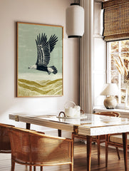 Illustrative linocut style print of an eagle flying over textured olive green hills under a, in a styled room mockup