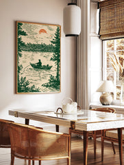 Illustrative print of a fisherman in a boat on a lake, framed by pine trees, under a setting sun, in a styled room mockup