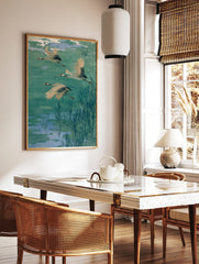 Three flying cranes with red heads and black wing tips over a teal and blue pond with reeds, in a styled room mockup