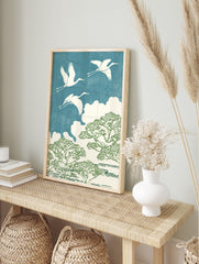 Three white cranes flying over green trees and white clouds against a textured blue sky in an, in an alternate framed mockup