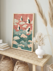Four white cranes with red crowns fly over teal waves and pink clouds against a coral sky in, in an alternate framed mockup