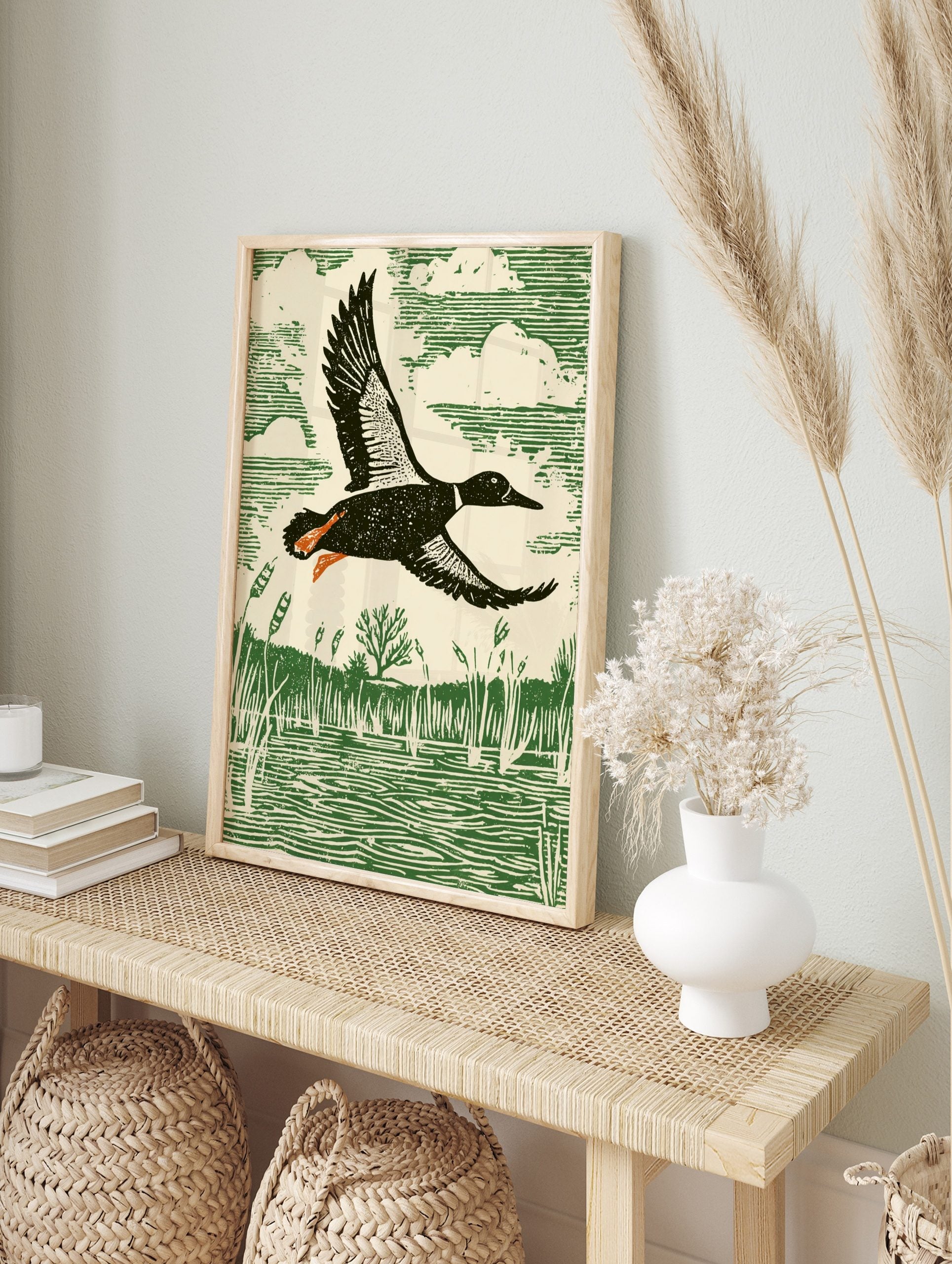 Linocut style print of a duck flying over a marsh with reeds and water, in green, cream, in an alternate framed mockup