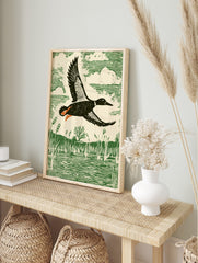 Linocut style print of a duck flying over a marsh with reeds and water, in green, cream, in an alternate framed mockup