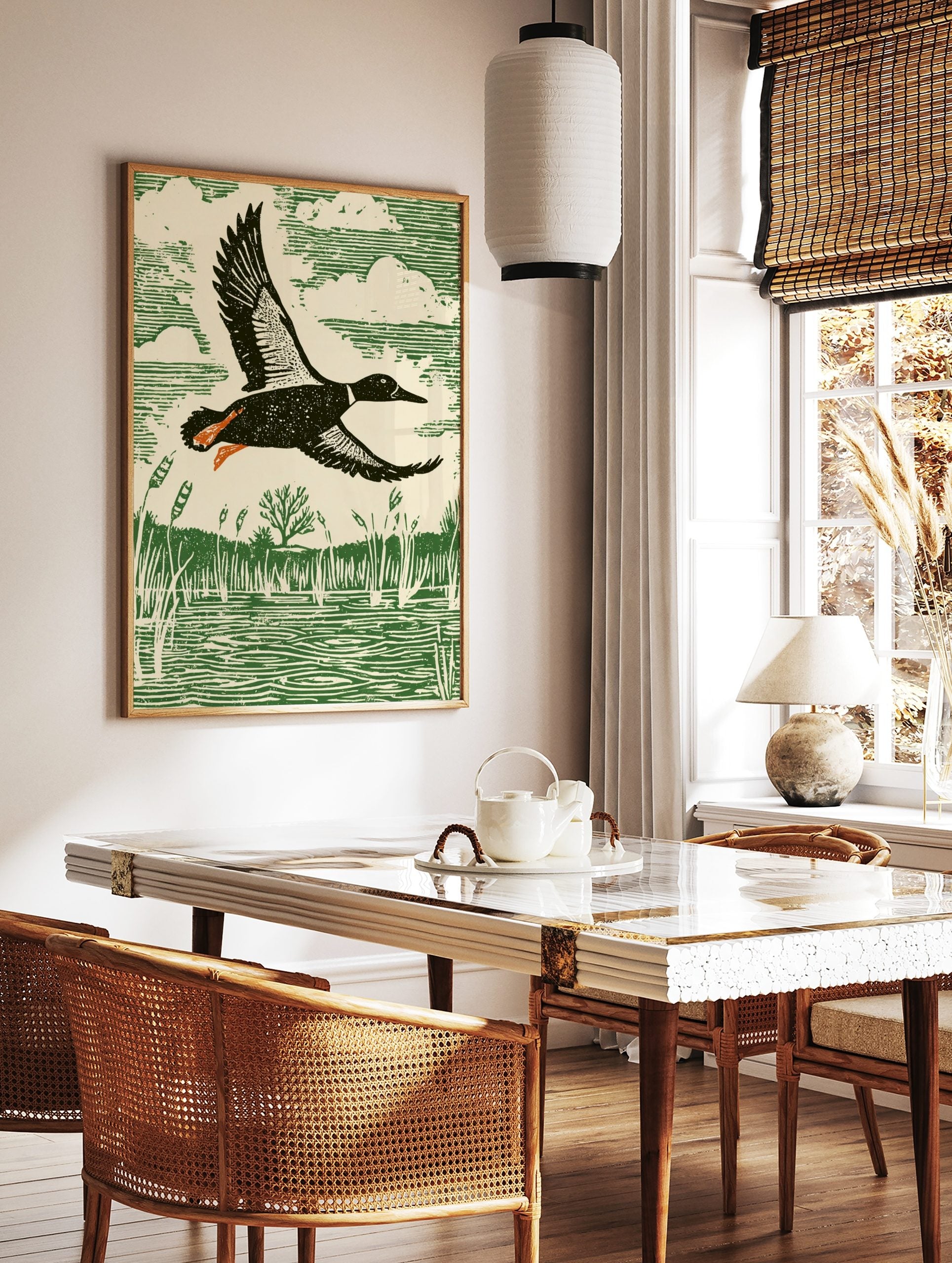 Linocut style print of a duck flying over a marsh with reeds and water, in green, cream, black, and, in a styled room mockup
