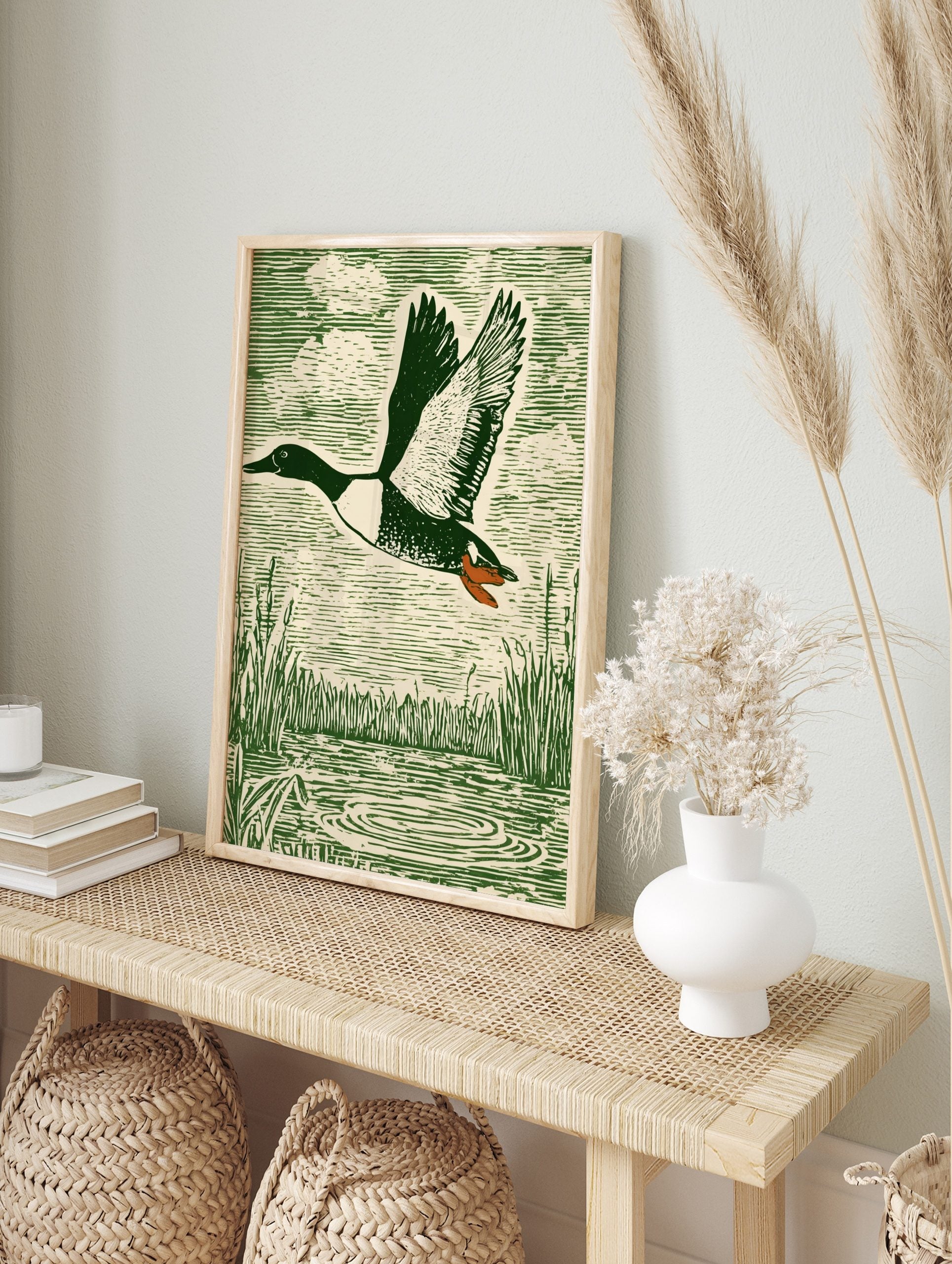 Green woodcut-style print of a flying duck with spread wings over water and reeds, in an alternate framed mockup